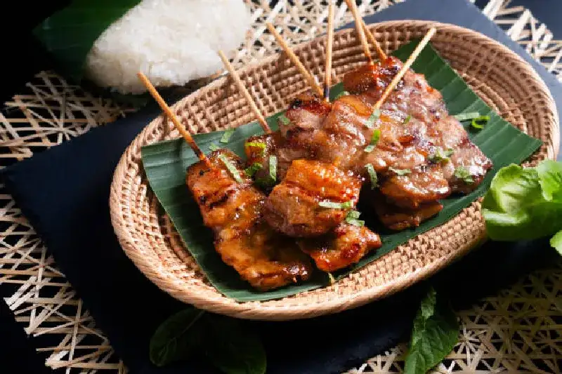 Thailändische Street-Food-Spezialität „Moo Ping“: gegrillte Schweinespieße serviert mit Reis, auf schwarzem Hintergrund mit Platz für Text.