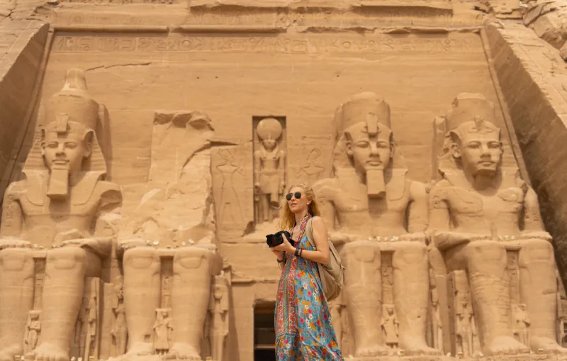 A traveler stands in front of the monumental rock-cut façade of Abu Simbel Temple, with the colossal statues of Pharaoh Ramesses II carved into the sandstone cliffs.