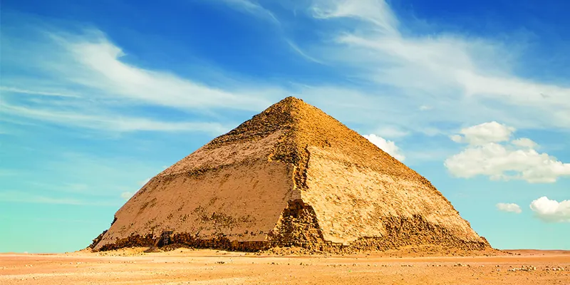 A stunning view of the Bent Pyramid at Dahshur, Egypt, with its unique angled structure standing under a sky filled with puffy clouds, highlighting the architectural ingenuity of ancient Egypt.