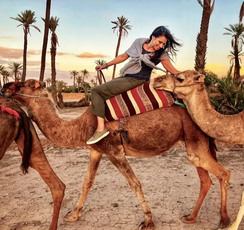 A smiling woman rides a camel at sunset in a desert outside of Marrakech, Morocco