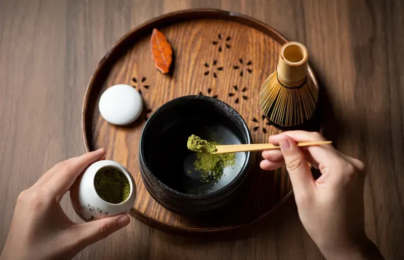 maos-adicionando-po-de-matcha-em-tigela-preta-com-colher-de-bambu-sobre-bandeja-de-madeira