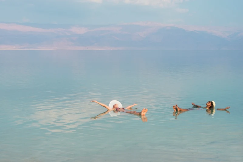 Two people floating on calm turquoise waters of the Dead Sea with distant mountains under partly cloudy sky.