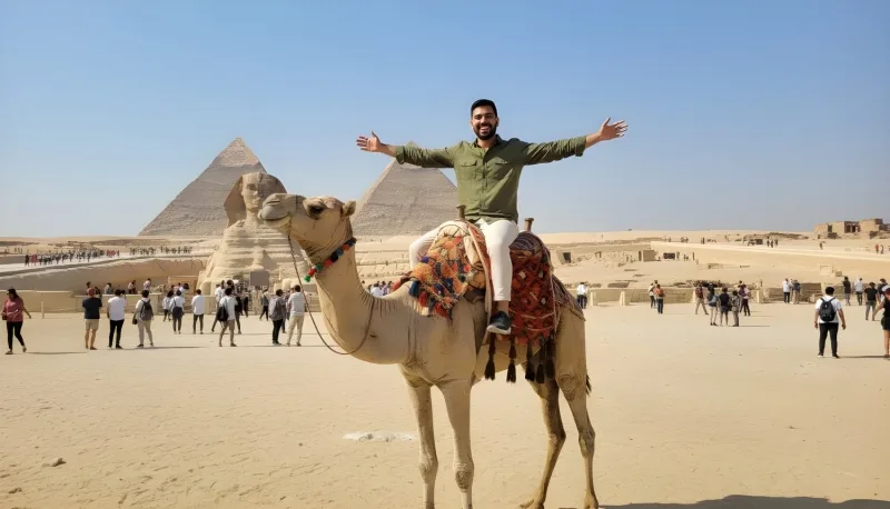 Happy male tourist riding a camel near the Great Sphinx and Pyramids of Giza, with other tourists walking in the background.