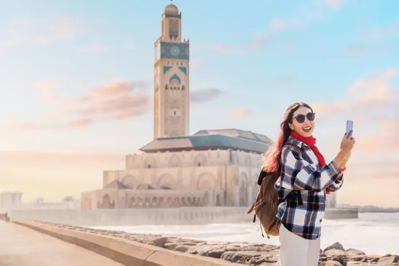 Turista sonriente capturando con su teléfono la impresionante Mezquita Hassan II al atardecer, rodeada del vibrante ambiente de Casablanca, Marruecos.