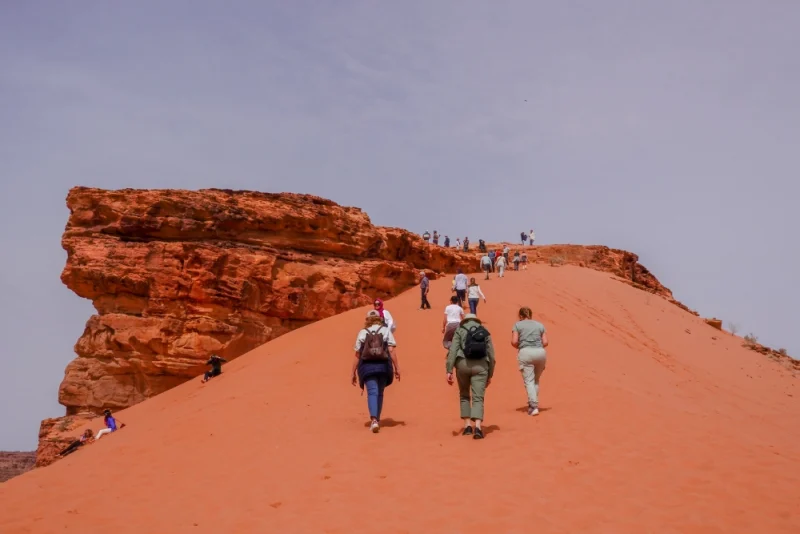 Group hiking up steep orange sand dune toward towering red sandstone formation under clear sky.