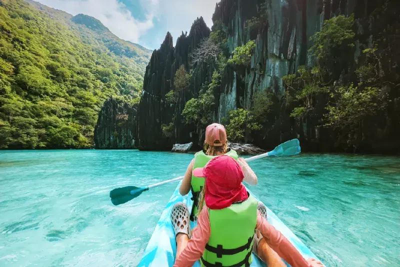 Two travelers kayaking through the emerald lagoon of El Nido, Palawan, with dramatic limestone formations and tropical scenery.