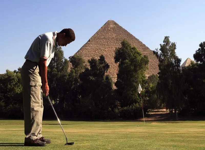 A golfer preparing to putt on a green with the Great Pyramid of Giza rising in the background at the Mena House Oberoi Golf Club.