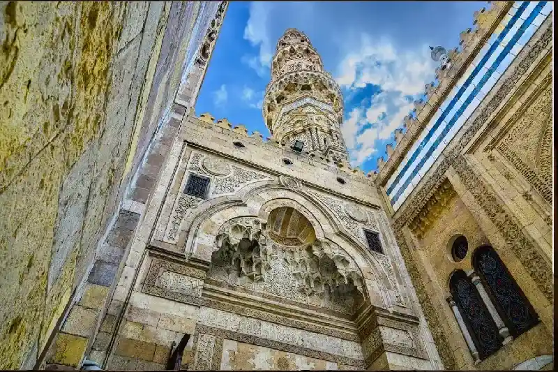 Al-Azhar-Moschee im alten Kairo, Ägypten. Untersicht eines alten Steinbaus mit kunstvollen Schnitzereien und einem hohen Minarett vor blauem Himmel mit Wolken.