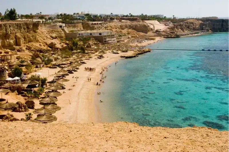 Luftaufnahme eines belebten Sandstrandes mit Strohhütten, türkisfarbenem Meer und Hotels im Hintergrund.