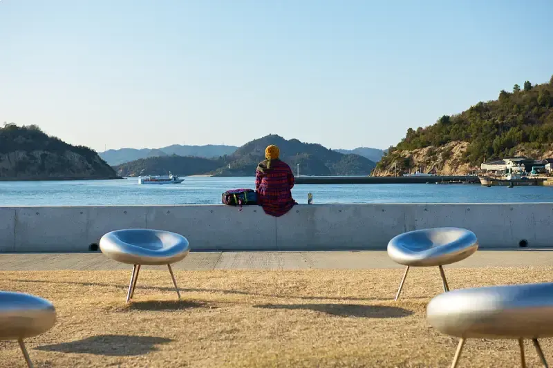  pessoa-sentada-de-costas-observando-o-mar-em-naoshima-japao-cercada-por-bancos-metalicos-em-formato-de-gota