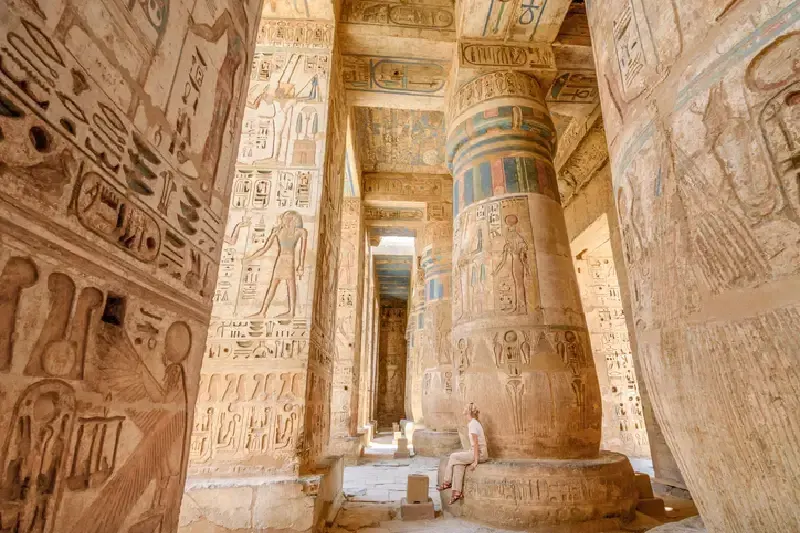 Frau sitzt auf einer Säule im Tempel von Ramses III. in Luxor, Ägypten, umgeben von antiken Ruinen und ägyptischer Architektur.