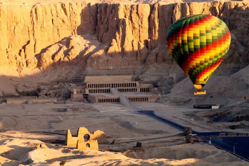 A colorful hot air balloon floats above the Mortuary Temple of Hatshepsut, set against the towering limestone cliffs of Luxor's West Bank at sunrise.