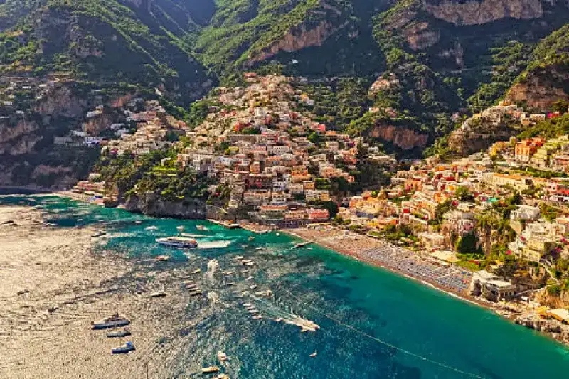 Bunte Häuser auf steilen Klippen entlang der Amalfi Coast, mit Blick auf das türkisfarbene Meer, enge Gassen und die charmante Küstenstadt Positano unter strahlendem Himmel.