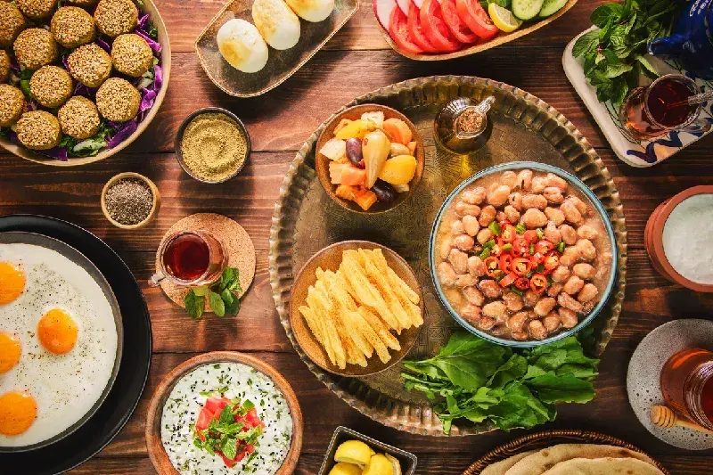 Ein reich gedeckter Tisch mit traditionellen ägyptischen Gerichten, darunter eine Schale Foul Medames mit Chilischoten, Falafel (Ta'ameya), Spiegeleier, frisches Gemüse, Fladenbrot und Gläser mit schwarzem Tee auf einem rustikalen Holzhintergrund.