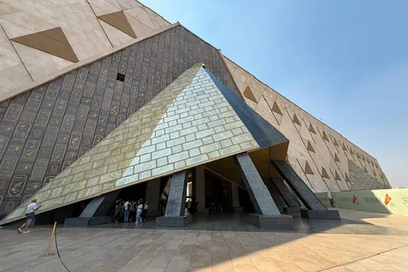 Haupteingang des Grand Egyptian Museum mit moderner, dreieckiger Architektur, Besuchern vor dem Gebäude und weitem Blick auf den Vorplatz in Giza.