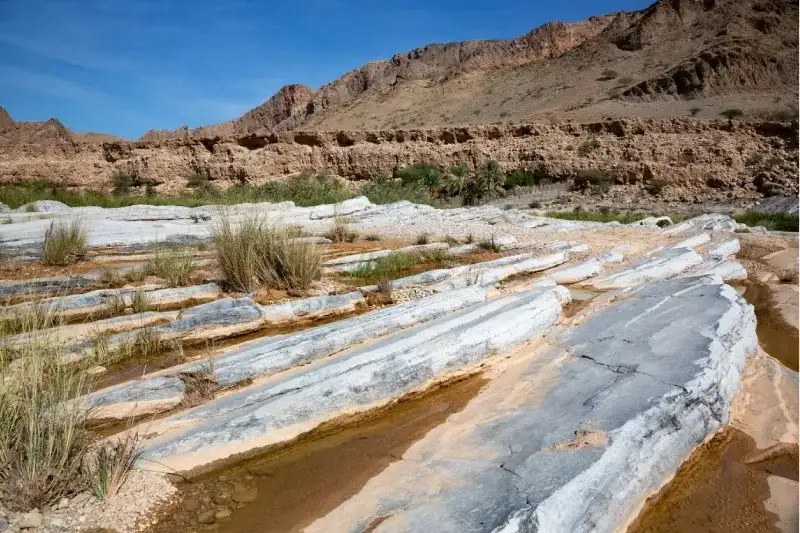 Formazioni rocciose  con piccole pozze d'acqua in wadi Wadi Al Arbaeen, con montagne sullo sfondo.