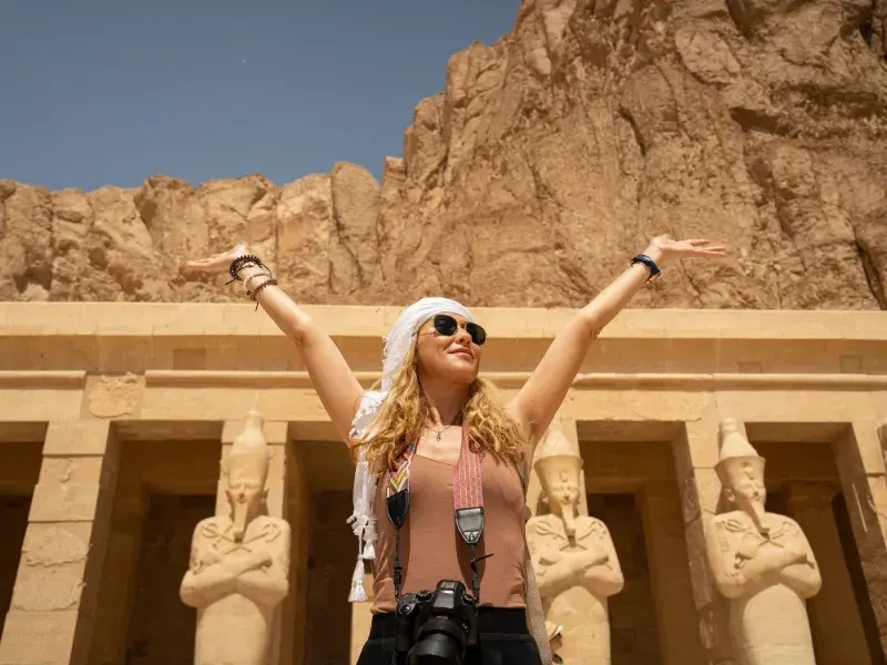 Donna a braccia aperte davanti al Tempio di Hatshepsut a Luxor, con grandi statue di faraoni e falesie rocciose.