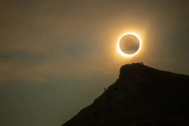 A striking total solar eclipse forms a radiant ring of fire above a rugged mountain peak.