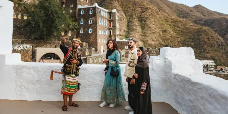 Guía turístico explicando la historia y riqueza cultural de Rijal Almaa en Abha Asir, rodeado de arquitectura tradicional y montañas.