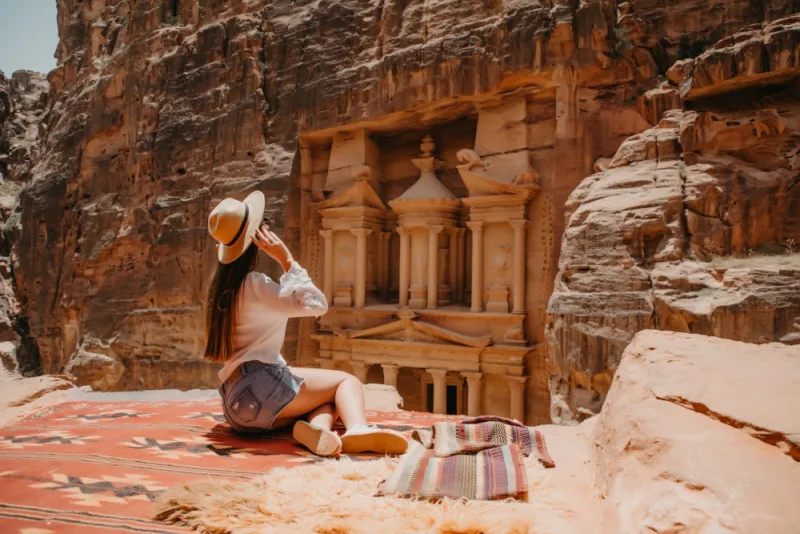 Person seated on rocky ledge overlooking Petra’s Treasury carved into sandstone cliffs.