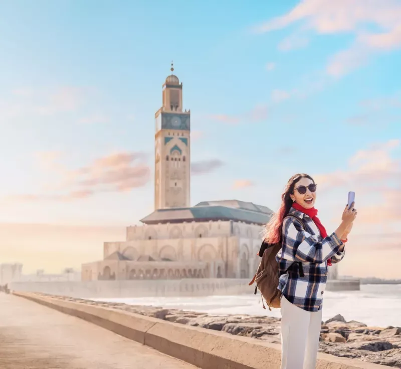 Smiling tourist capturing the stunning Hassan II Mosque by phone at sunset, surrounded by the vibrant atmosphere of Casablanca, Morocco