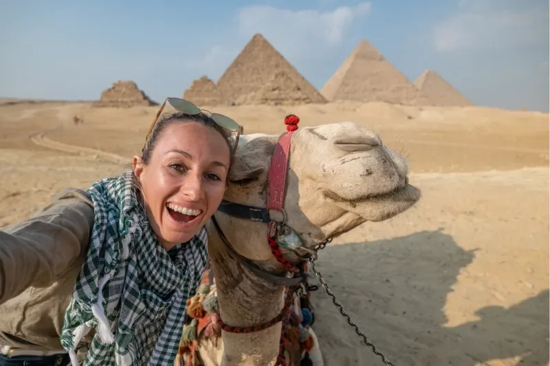 Vista frontale di una turista che visita le Piramidi di Giza in Egitto. Un cammello è al suo fianco.