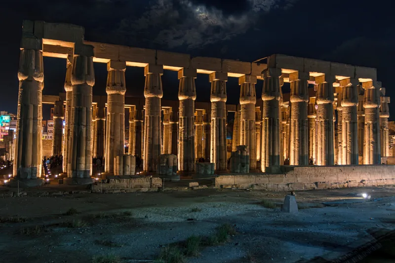 Turistas visitando el templo de Luxor por la noche con la luna de fondo.
