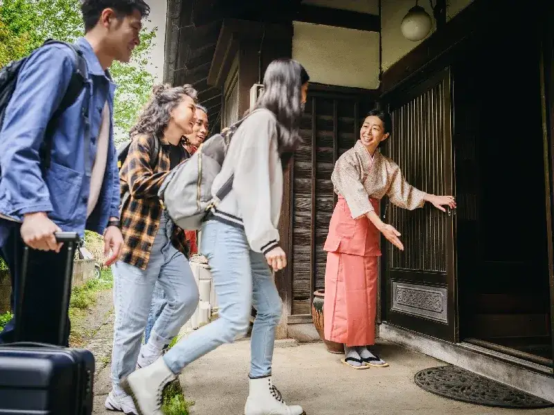 Mujeres japonesas dan la bienvenida a cuatro mochileros con equipaje, les abren la puerta y los hacen pasar al interior del edificio.