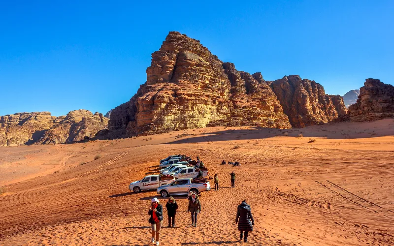 4x4 vehicles waiting for tourists who descend from rocks on top of red dunes of Wadi Rum desert