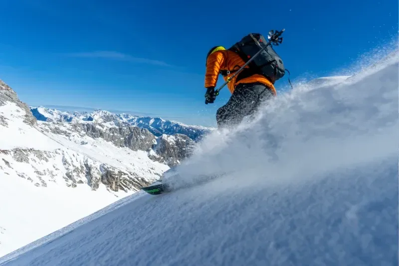 Skibergsteiger in orangefarbener Ausrüstung fährt durch frischen Pulverschnee auf einer steilen Alpenpiste.