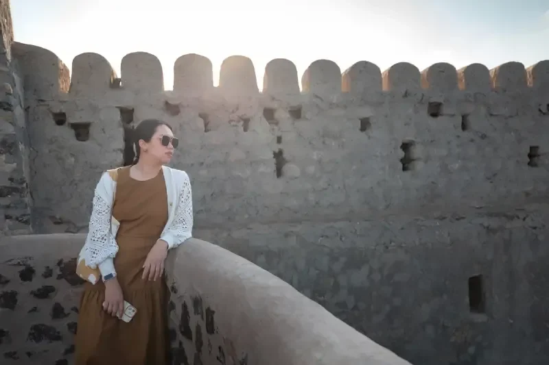 Una donna elegante posa davanti a un'antica fortezza di Muscat, in Oman, tra fascino storico e stile contemporaneo.