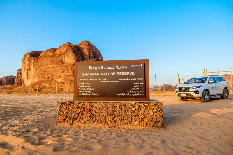 Cartel de bienvenida a la Reserva Natural de Sharaan en AlUla, Arabia Saudita, rodeado de formaciones de arenisca y un SUV blanco.