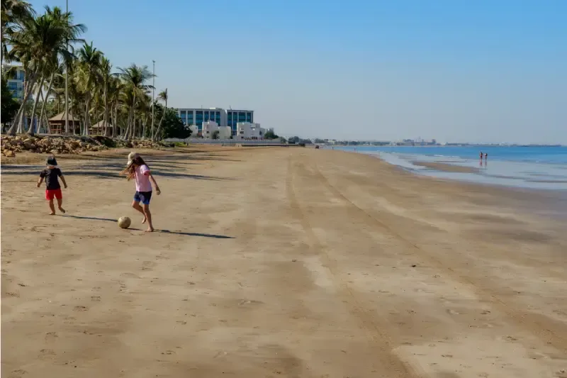 Due bambini giocano sulla spiaggia di Al Qurum a Muscat, tra palme, acque cristalline e un'atmosfera ideale per una vacanza in famiglia.