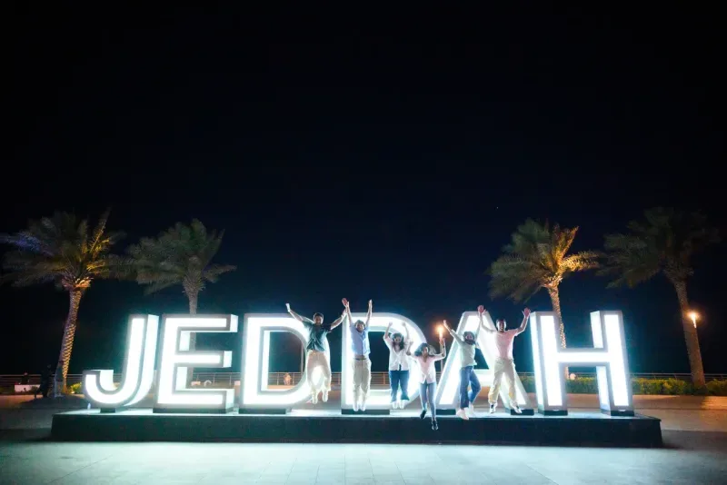 Friends celebrating beside the glowing Jeddah sign at night.