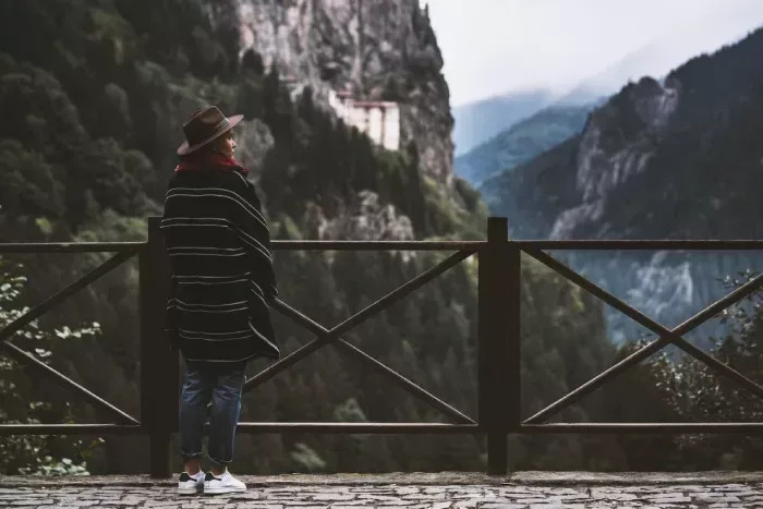 Young woman wearing a cape with a cowboy hat, standing in front of the wooden fences. Watching Sumela Monastery and the mountains over the hill