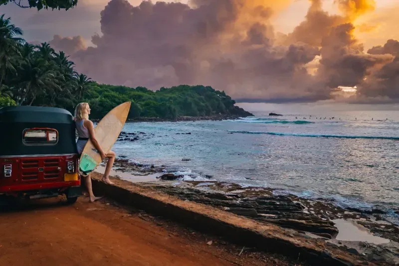 Una surfista está de pie junto a su tuk-tuk, contemplando el océano al atardecer con una tabla de surf a su lado.