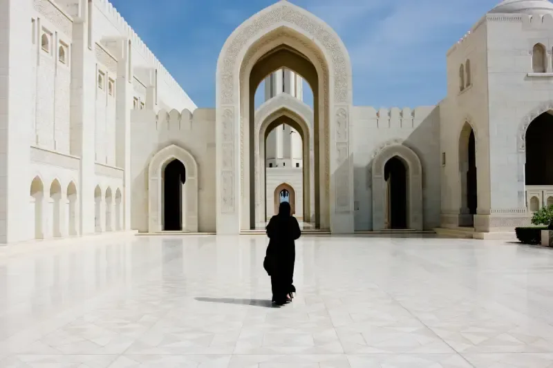 Donna in abaya nera che cammina verso il grande arco della Grande Moschea del Sultano Qaboos a Muscat, in Oman.