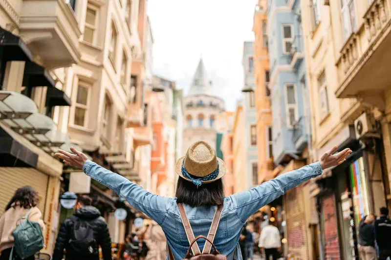 Woman with a backpack and sun hat raising her arms on a colorful street leading to the Galata Tower in Istanbul, Turkey
