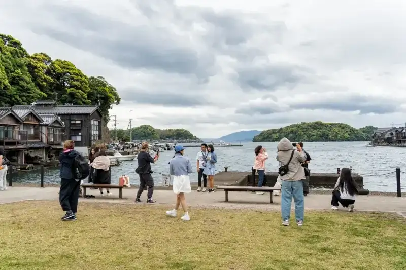 uristas-observando-a-baia-em-ine-no-funaya-no-japao-vila-costeira-com-casas-barco-tradicionais-a-beira-mar-sob-um-ceu-nublado