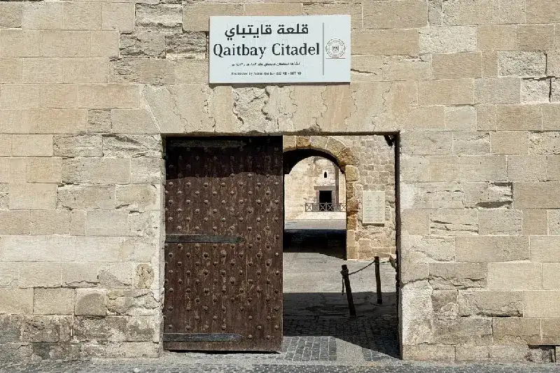Das Eingangstor der Zitadelle von Qaitbay in Alexandria, Ägypten, mit einem offiziellen Schild an einer historischen Steinmauer.