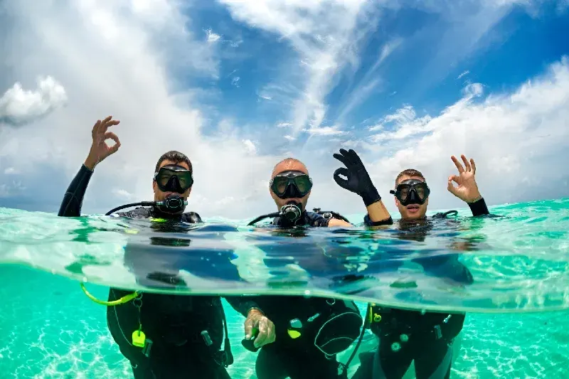 Drei Taucher im flachen türkisfarbenen Wasser mit Tauchausrüstung, zwei zeigen das OK-Zeichen unter einem sonnigen Himmel.