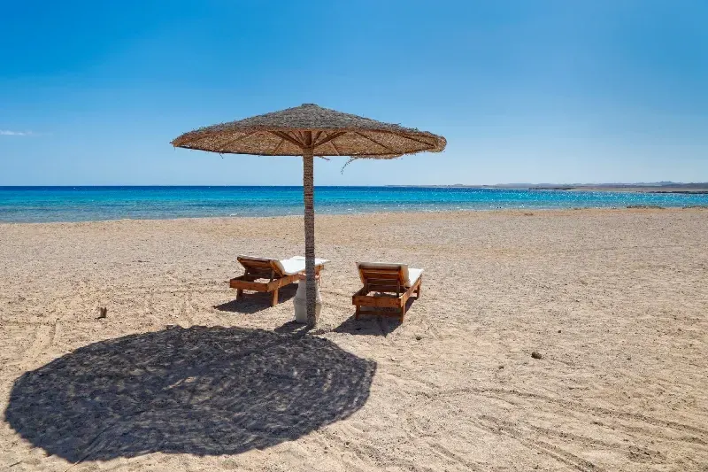 Strohschirm mit zwei Liegestühlen am sonnigen Sandstrand vor dem ruhigen blauen Meer unter klarem Himmel.