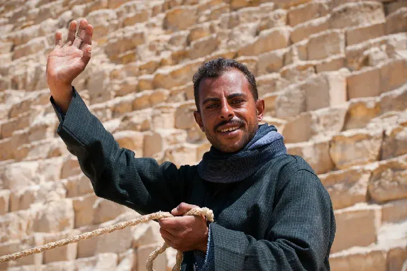 Ein ägyptischer Mann, der auf einem Kamel sitzt, winkt vor den Pyramiden von Gizeh in Ägypten zum Gruß.