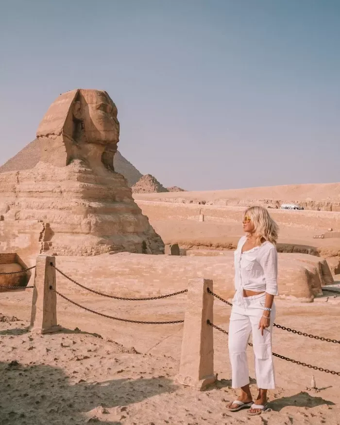 Ablonde lady in white shirt and pants stands infront of great sphinx