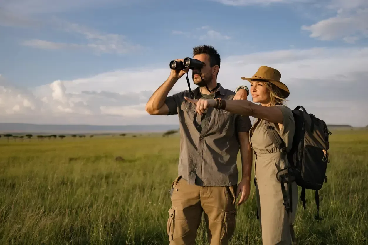 A couple on safari stands in the open savannah, one scanning the horizon with binoculars while the other points ahead, both immersed in the thrill of wildlife spotting.