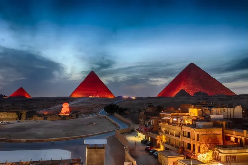 Die drei Pyramiden von Gizeh und die Sphinx rot angestrahlt während der Ton-und-Licht-Show, davor die Dächer von Gizeh