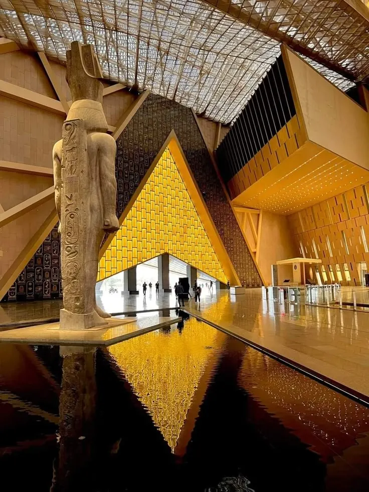 A statue of Ramesses II stands beside a reflecting pool inside the Grand Egyptian Museum, with modern geometric architecture and golden lighting dominating the background.