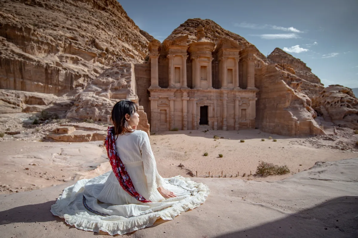 Person in white dress and red scarf seated before Petra’s Monastery carved into sandstone cliffs.