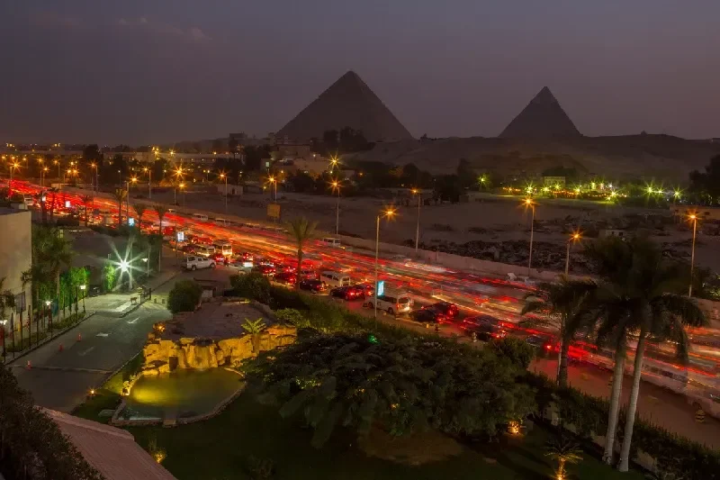  Verkehr in Kairo bei Nacht mit Blick auf die beleuchteten Pyramiden im Hintergrund.