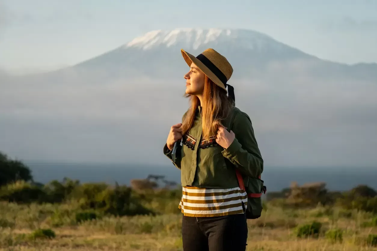 A woman stands peacefully in front of Mount Kilimanjaro, soaking in the view with a content smile and a backpack ready for adventure.